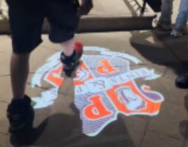 Video: orange restaurant crest gobo shines on a night sidewalk as pedestrians walk past