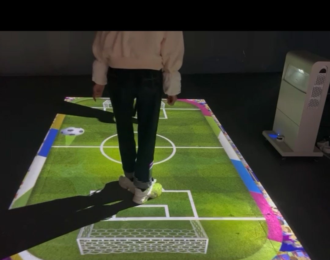 Video: kid kicks a virtual soccer ball across a glowing green pitch projected on the floor of a dark play space
