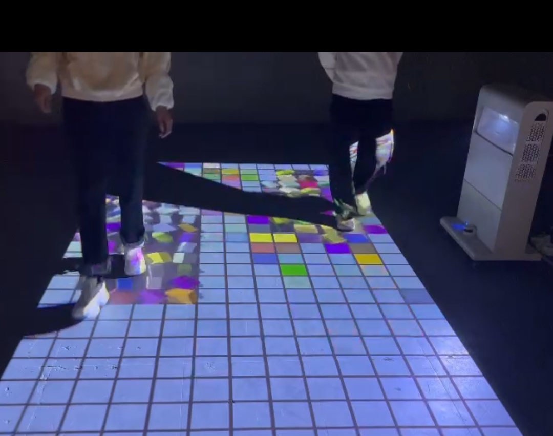 Video: two kids reveal colorful mosaic tiles by walking on a white grid projected across a dark room floor