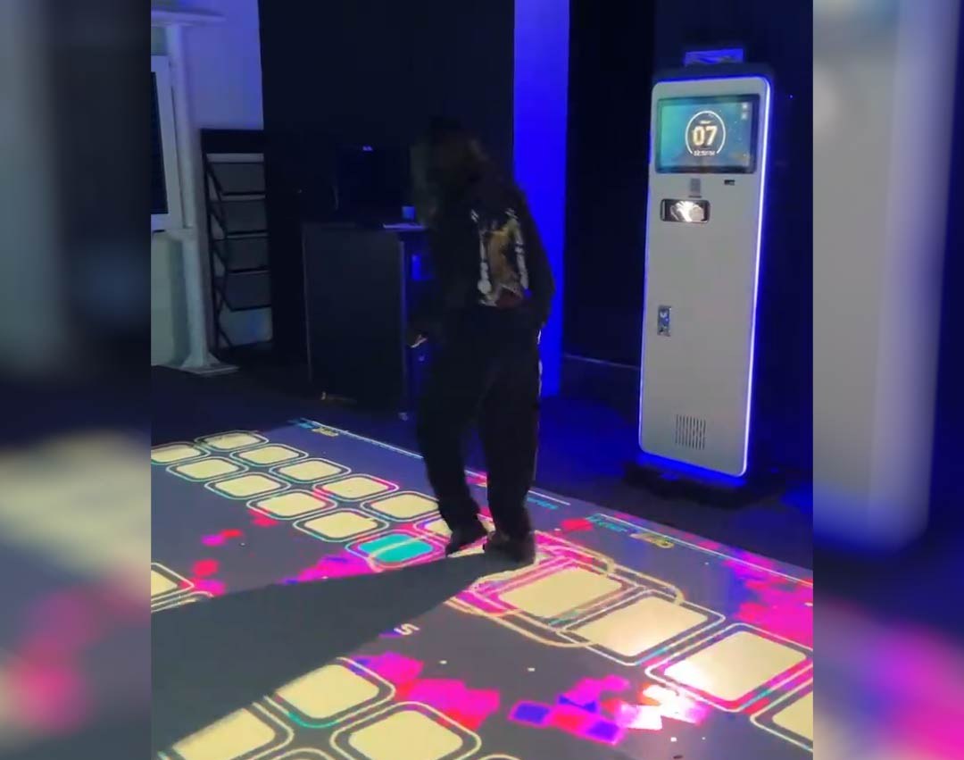 Video: dancer hops between golden light-up squares projected on a game-room floor beside a countdown kiosk
