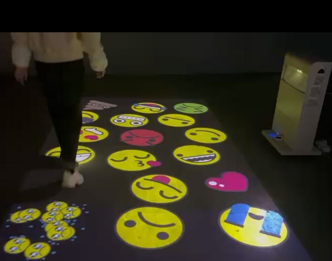 Video: child walks through giant smiley-face emojis projected on a dark playroom floor from a mobile tower projector
