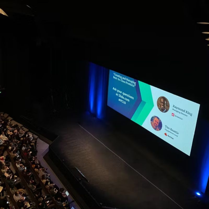 Wide LED video wall displaying a conference session slide above a stage in a packed auditorium — Infinity Projectors