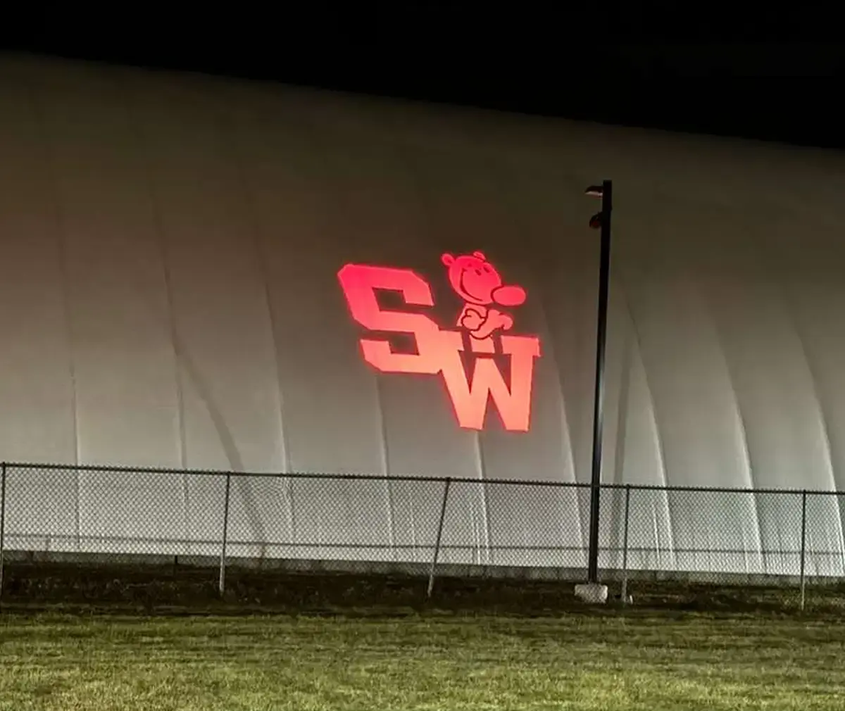Red school initials with a cartoon mascot beamed onto a white sports dome behind a chain-link fence — Infinity Projectors