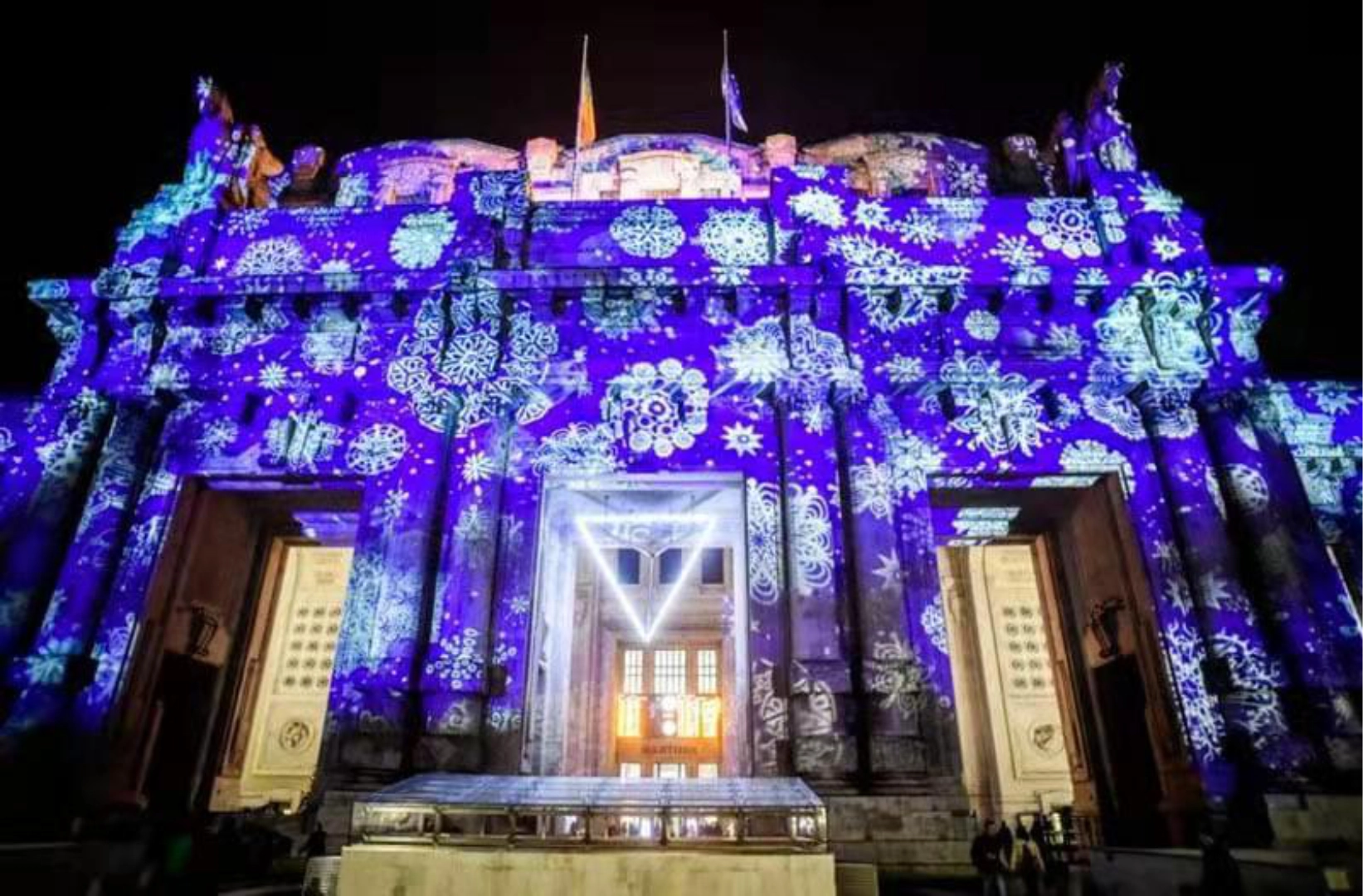 Snowflake patterns blanket a grand columned building in deep blue light during a night projection show