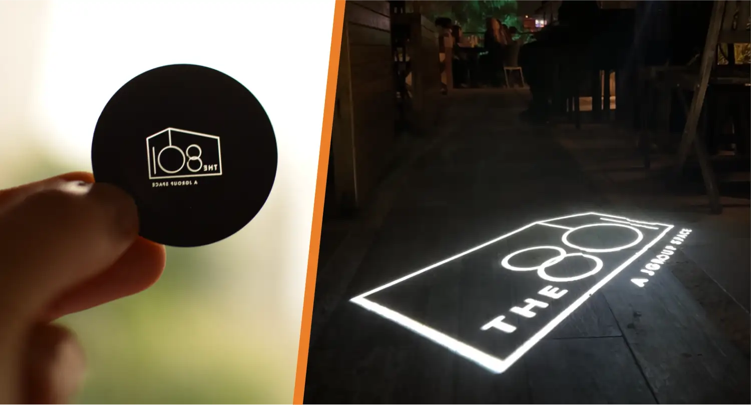 Hand holding a custom gobo disc beside its white logo projected on a wooden patio floor at night