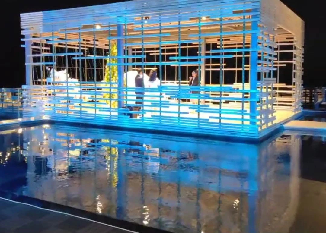 Video: rippling blue light bathes a slatted poolside pavilion, reflections shimmering on the water at night