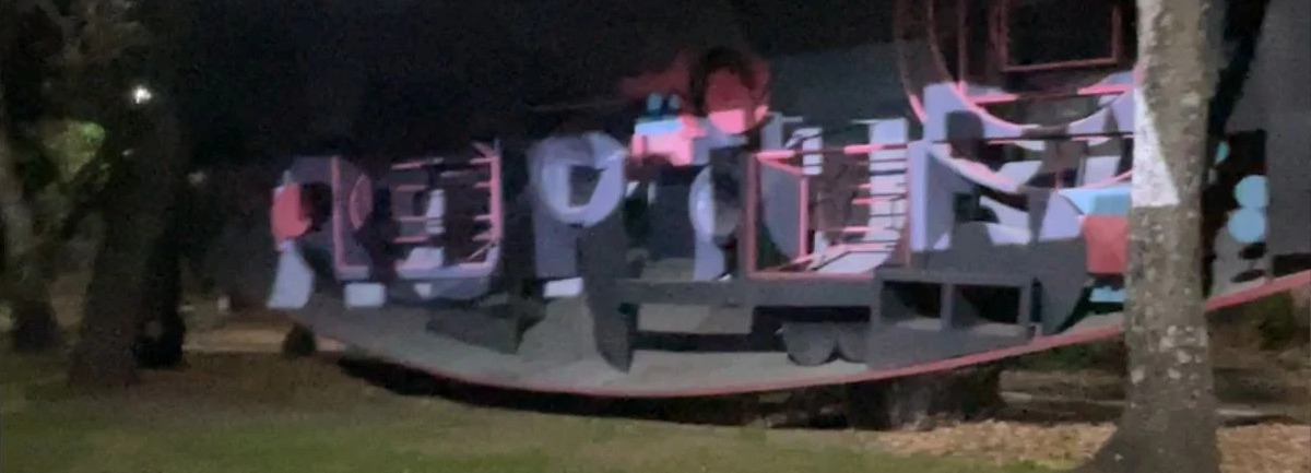 The word RUPTURA glows across a park art installation at night, cast by a high wattage gobo projector; branded image