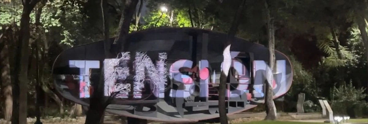 TENSION in tall letters projected onto a curved outdoor pavilion among trees at night; high wattage gobo projector