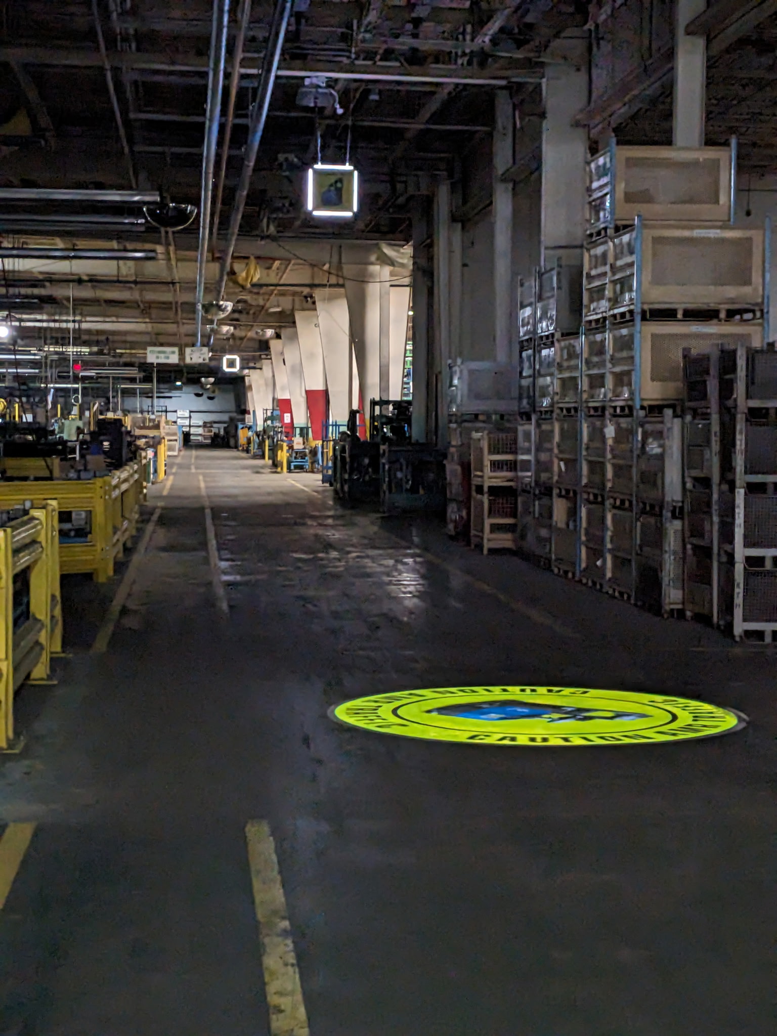 Caution safety graphic floor projection inside a warehouse and factory aisle at KTH Parts Industries, with pallet racking and shipping containers visible — Infinity Projectors