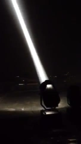 Single searchlight beam shooting into the night sky from a rooftop, distant city lights on the horizon — outdoor sky searchlight in action