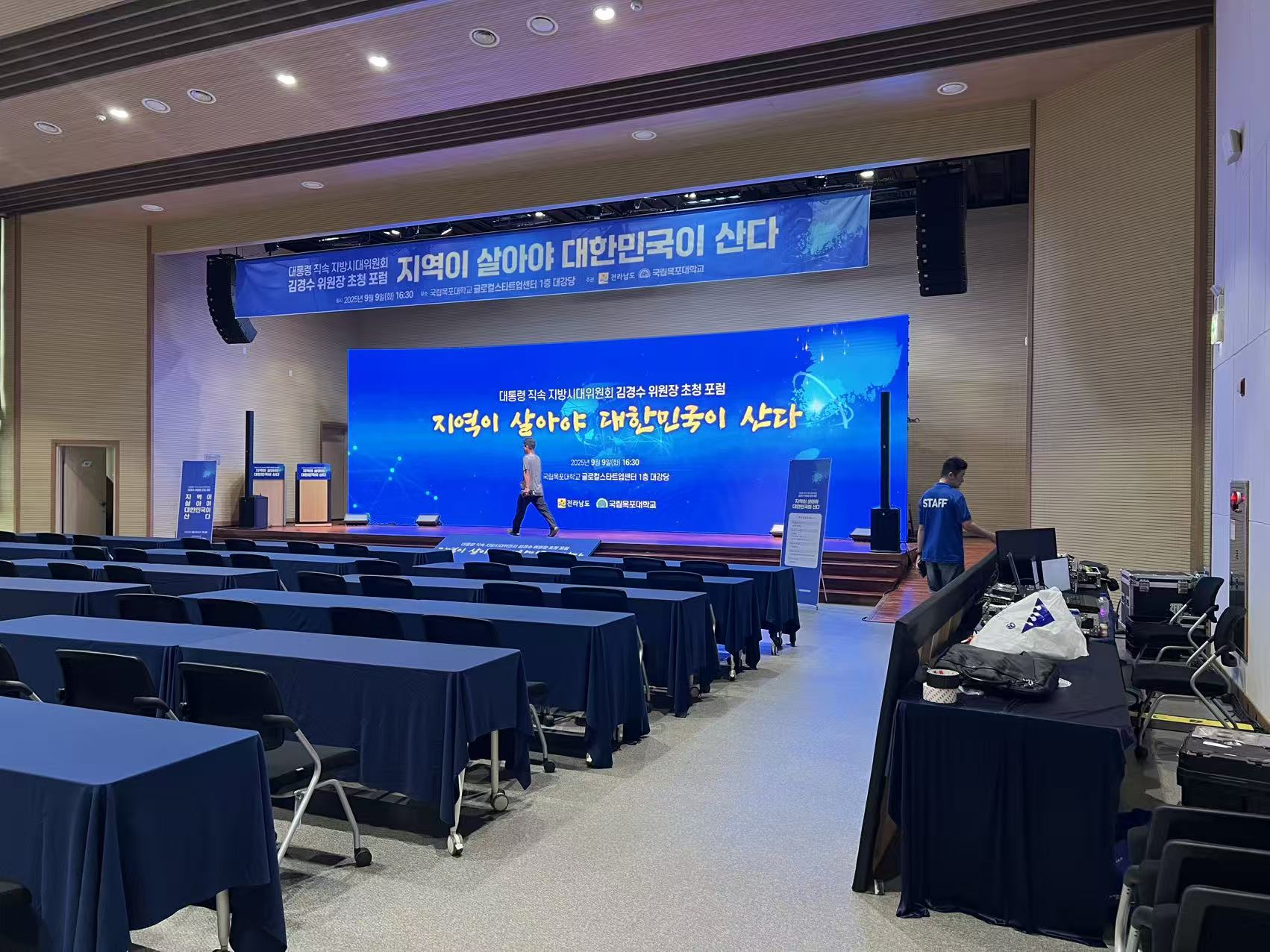 University auditorium with a blue LED backdrop and hanging banner for a Korean forum, rows of navy-draped tables in front — Infinity Projectors