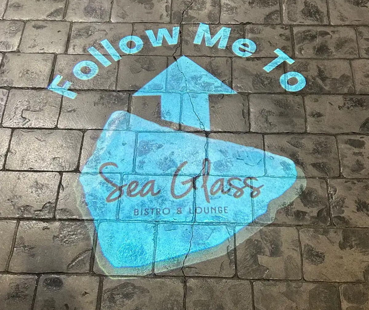 Teal 'Follow me to' arrow and bistro logo gobo lighting the way across stamped concrete pavers — real install