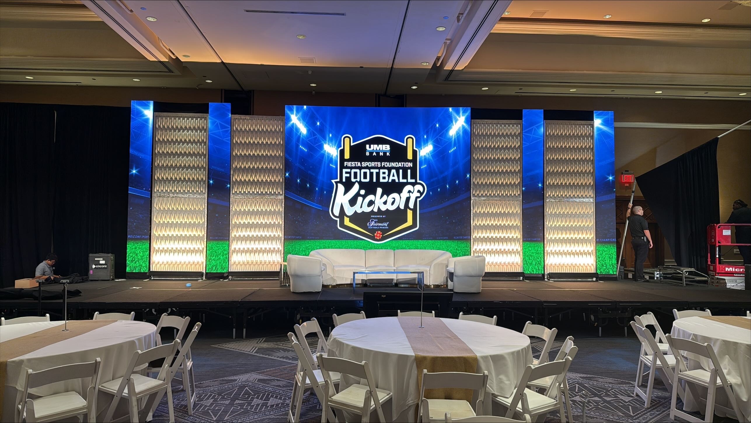 Hotel ballroom stage with an LED video wall showing a stadium-themed football kickoff graphic behind white lounge seating