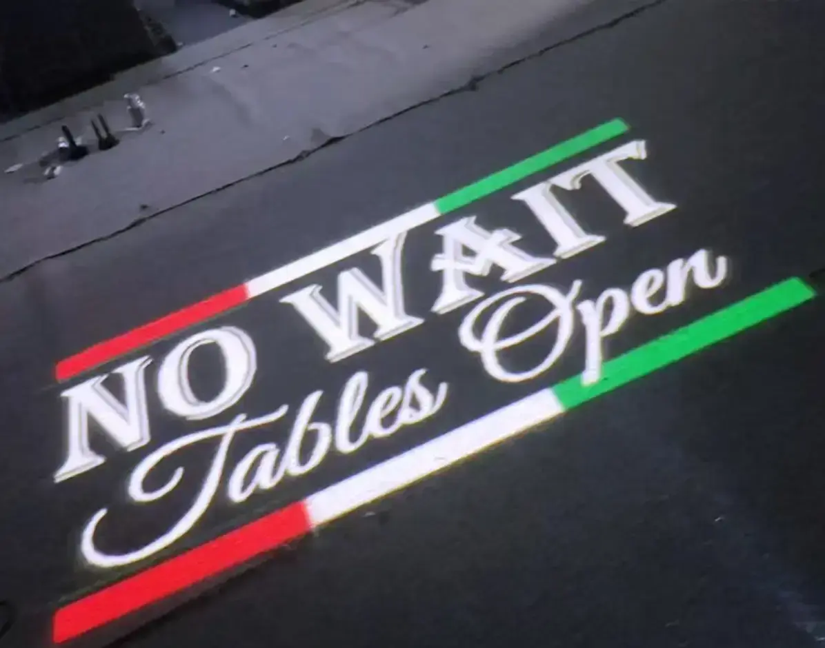 'No Wait, Tables Open' script in Italian-flag stripes projected on dark pavement outside a restaurant — real install