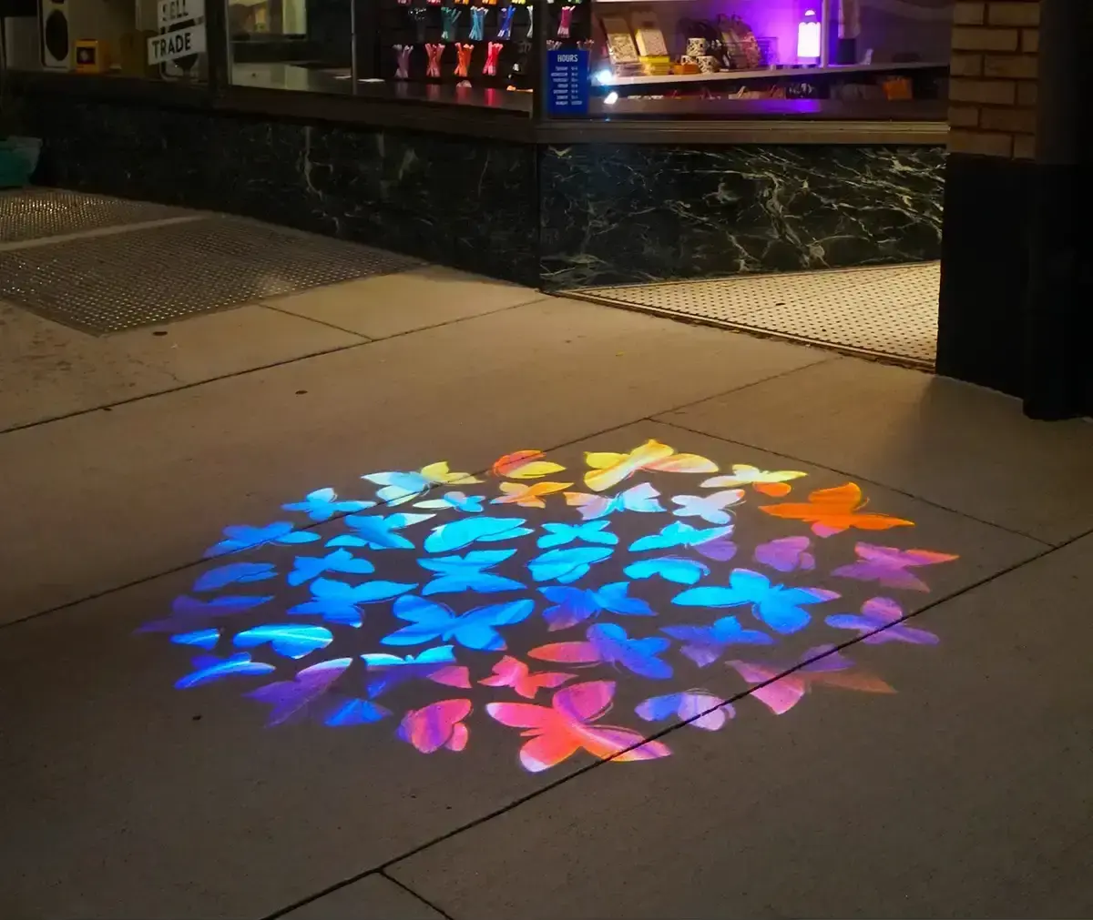 Swarm of blue, pink, and orange butterflies projected on the pavement outside a storefront at night — Infinity Projectors