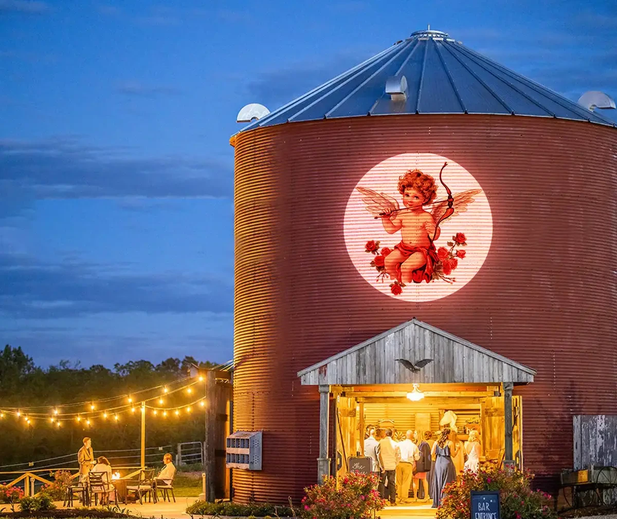 Red cupid-and-roses gobo glowing on a rustic silo venue at dusk as wedding guests gather under string lights — Infinity Projectors