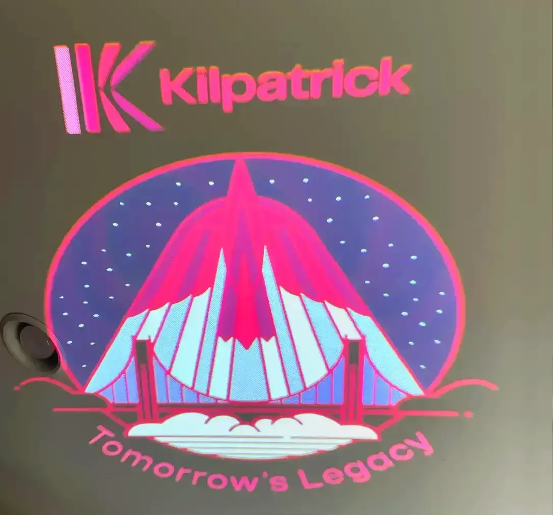 Hot-pink corporate crest with bridge artwork test-projected on a wall, gobo projector lens visible at left — Infinity Projectors