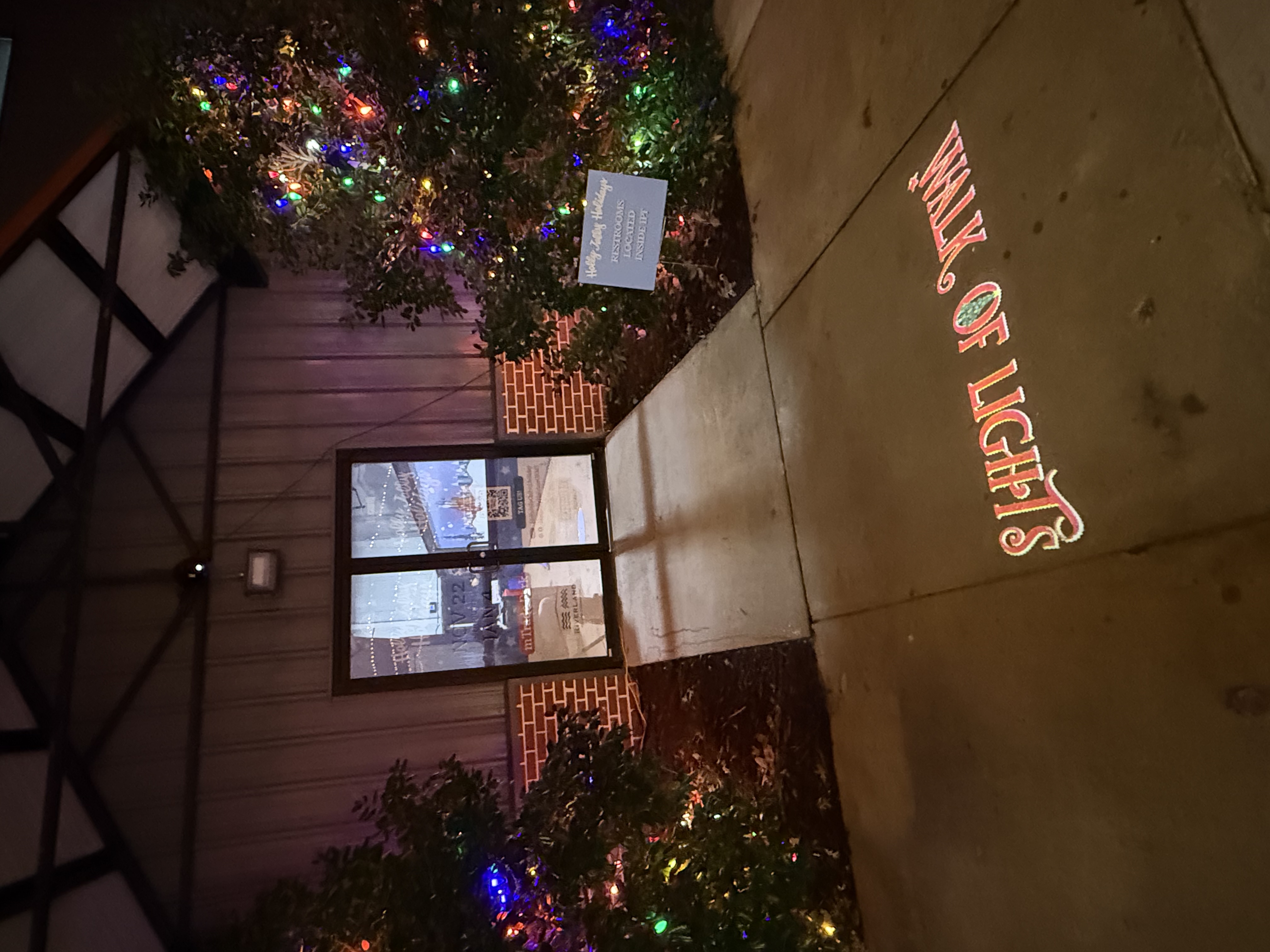 Sideways photo of red Walk of Lights lettering projected on a sidewalk beside holiday-lit shrubs